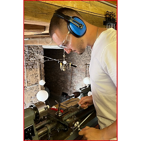 Mann mit Gehörschutz und Schutzbrille an der Drehbank. Er arbeitet an Metall, der Hintergrund zeigt einen Steinbogen.